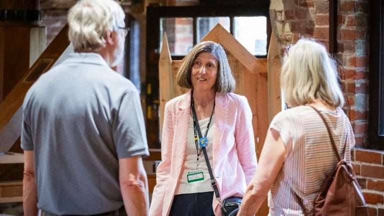 A person wearing a light pink jacket and a lanyard stands indoors in front of a wooden structure, speaking with two other people who are facing toward the camera. The setting appears to be a brick-walled interior space with warm lighting and visible wooden architectural elements.
